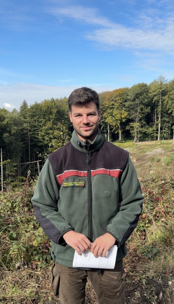 Ein Portrait von Förster Rico Hollenberg am Rand einer freien Fläche mit Wald im Hintergrund