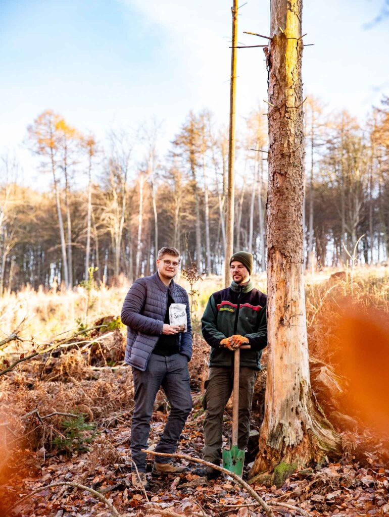 Julian Meyer, von der Werbeagentur die medienvilla, und Förster Rico Hollenberg stehen an einer toten Fichte im Wiehengebirge mit einer Gebäcktüte von der Bäckerei Schmidt