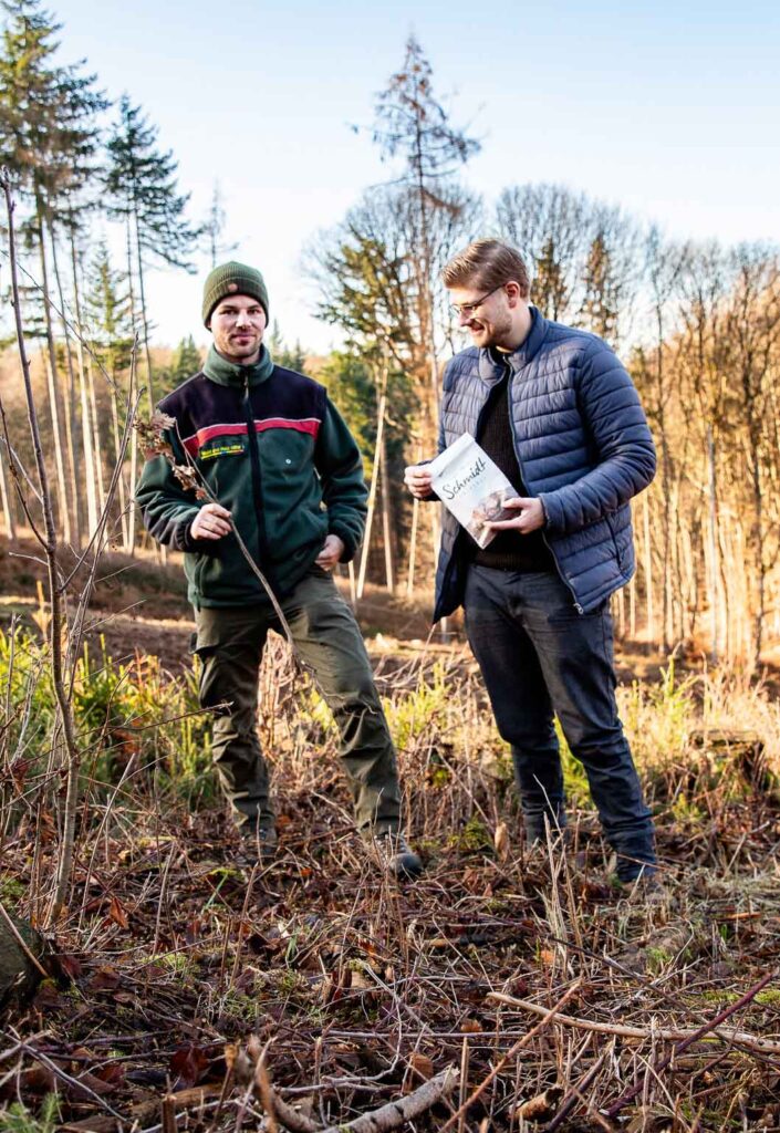 Förster Rico Hollenberg und Mediendesigner Julian Meyer stehen neben einander im Wiehengebirge. Rico Hollenberg hält einen Baum Setzling und Julian Meyer eine Bäckerei Schmidt Tüte